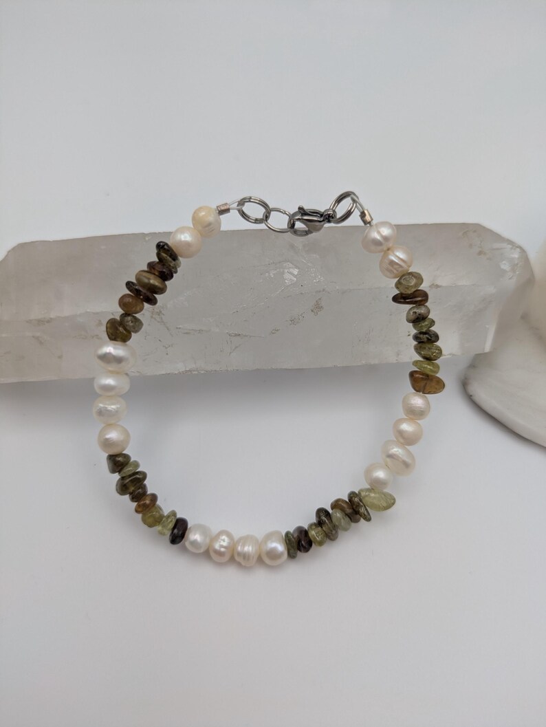 K&ouml;nnte beinhalten: Ein Armband aus wei&szlig;en Perlen und gr&uuml;nen und braunen Edelsteinperlen. Das Armband hat einen silbernen Verschluss.