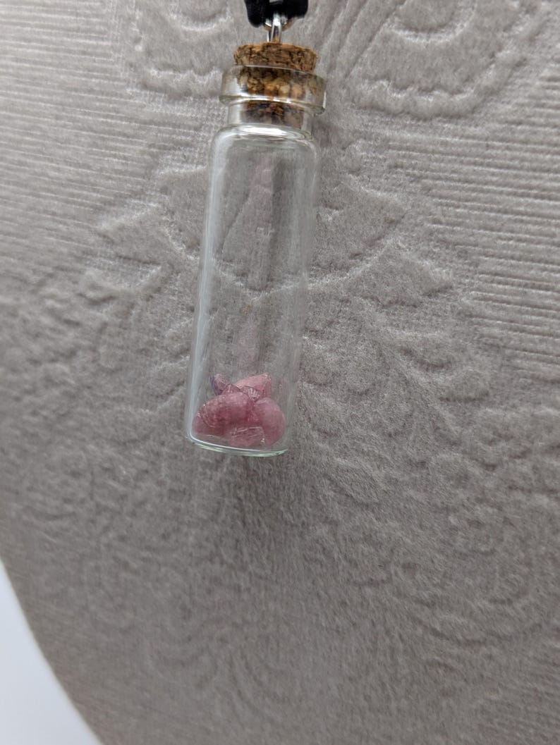 Puede incluir: Un collar con una peque&ntilde;a botella de vidrio con un tap&oacute;n de corcho. La botella contiene cristales rosas.