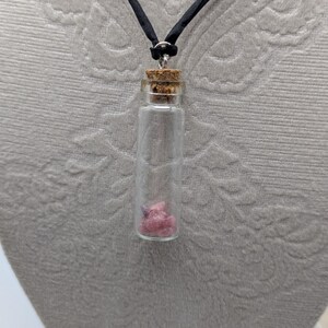 Puede incluir: Un collar con un vial de vidrio y un tap&oacute;n de corcho, en un cord&oacute;n negro. El vial contiene peque&ntilde;os cristales rosas y morados.