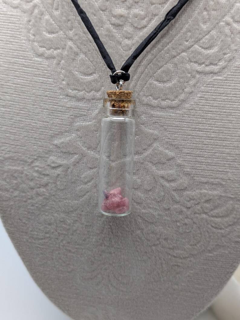 Puede incluir: Un collar con un vial de vidrio y un tap&oacute;n de corcho, en un cord&oacute;n negro. El vial contiene peque&ntilde;os cristales rosas y morados.