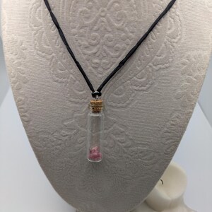Puede incluir: Un collar de cord&oacute;n negro con un peque&ntilde;o colgante de vidrio en forma de frasco que contiene un cristal rosa. El frasco tiene un tap&oacute;n de corcho.