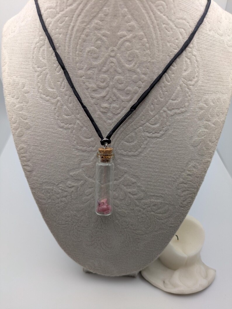 Puede incluir: Un collar de cord&oacute;n negro con un peque&ntilde;o colgante de vidrio en forma de frasco que contiene un cristal rosa. El frasco tiene un tap&oacute;n de corcho.