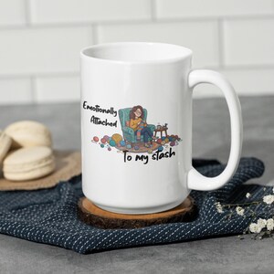 Può includere: Tazza in ceramica bianca con un manico grande, con la scritta "Emotionally Attached To my stash" e un'illustrazione di una persona che lavora a maglia. La tazza è appoggiata su un sottobicchiere di legno e un panno blu fantasia.