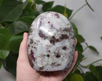 May include: A white, gray, and red polished crystal with a rounded shape. The crystal has a speckled appearance with red and gray inclusions.