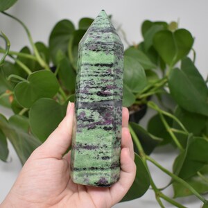 May include: A green, black, and pink striped crystal point. The crystal is about 6 inches tall and is held in a hand.