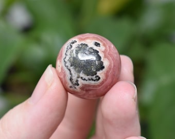 May include: A pink and black sphere with a swirling pattern. The sphere is made of a natural stone and has a smooth, polished surface.