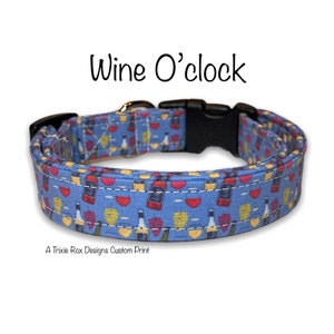 May include: A blue dog collar with a custom print of wine bottles and glasses. The collar has a black buckle and the text "Wine O'clock" is printed above the collar. The text "A Trixie Rox Designs Custom Print" is printed below the collar.