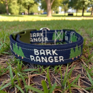 May include: A navy blue dog collar with the text "BARK RANGER" in white. The collar features a green and white forest pattern and a green plastic buckle. The collar is lying on green grass.