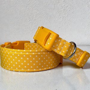 May include: A yellow dog collar with white polka dots and a matching yellow plastic buckle. The collar is made of fabric and has a metal D-ring for attaching a leash. The collar is designed for pets.