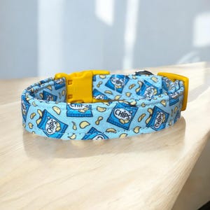May include: A light blue dog collar with a repeating pattern of chip bags and chips. The collar has a yellow plastic buckle. The word "CHIPS" is printed on the chip bags. The collar is sitting on a wooden surface.