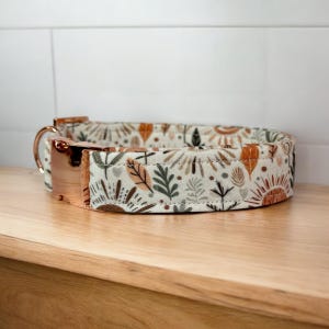 Pode incluir: Coleira para cães com um padrão floral em tons de castanho, verde e laranja sobre um fundo creme. A coleira tem uma fivela e um anel em D de metal em ouro rosa. A coleira está sobre uma superfície de madeira.