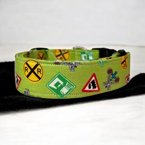 May include: A lime green dog collar featuring a repeating pattern of railroad crossing signs, signals, and symbols. The collar has a black plastic buckle and a metal D-ring for leash attachment. The fabric is a bright, eye-catching green.