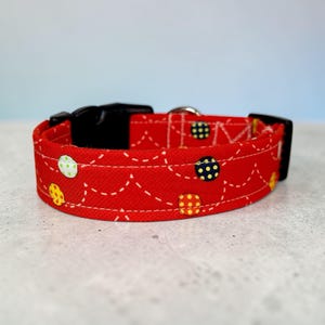 May include: A red dog collar with a black plastic buckle and a silver metal loop. The collar is decorated with a pattern of colorful polka dots and white stitching.