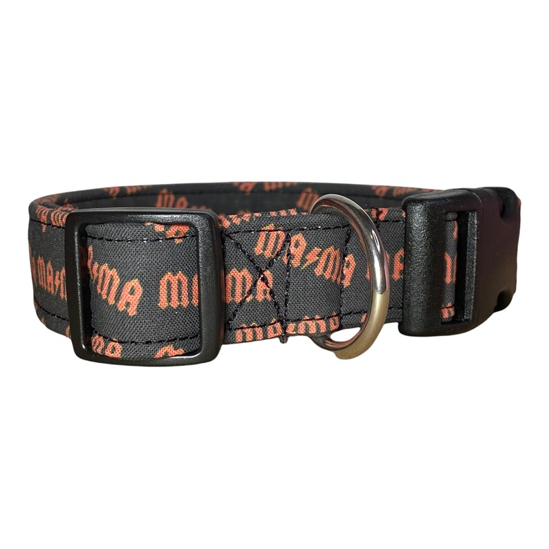 May include: Black dog collar with a silver D-ring and a black plastic buckle. The collar is made of fabric and has a repeating pattern of the word "mama" in orange.
