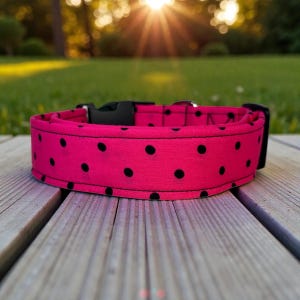 May include: A bright pink dog collar with black polka dots. The collar has a black plastic buckle and a metal loop for attaching a leash. The collar is sitting on a wooden surface.