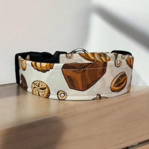 May include: A white dog collar with a black buckle and a pattern of brown and tan pastries, including donuts, croissants, and bread.