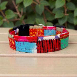 May include: A colorful dog collar with a patchwork design. The collar features a black plastic buckle and a metal loop. The fabric is patterned with red, blue, green, and yellow patches. The collar is sitting on a light brown surface.