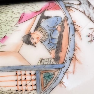 Puede incluir: Un plato de porcelana pintado a mano que representa una escena de una persona con una t&uacute;nica azul sentada en un barco. El barco est&aacute; rodeado de pinceladas verdes y marrones, lo que sugiere un entorno natural. El plato tiene un fondo blanco y un borde negro.