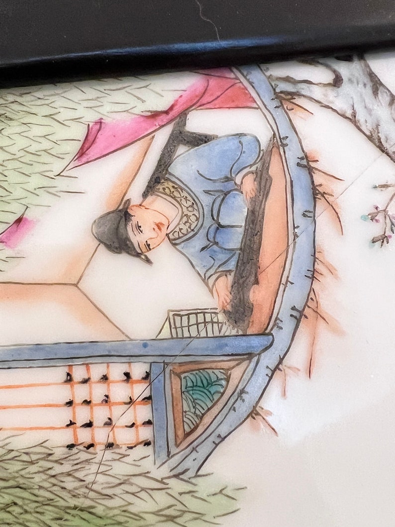 Puede incluir: Un plato de porcelana pintado a mano que representa una escena de una persona con una t&uacute;nica azul sentada en un barco. El barco est&aacute; rodeado de pinceladas verdes y marrones, lo que sugiere un entorno natural. El plato tiene un fondo blanco y un borde negro.