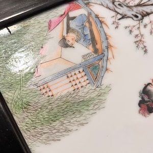 Puede incluir: Primer plano de un plato de porcelana pintado a mano que representa una escena de una persona en un barco con un &aacute;rbol y flores en el fondo. El plato est&aacute; enmarcado en madera negra.