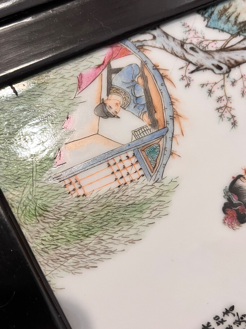 Puede incluir: Primer plano de un plato de porcelana pintado a mano que representa una escena de una persona en un barco con un &aacute;rbol y flores en el fondo. El plato est&aacute; enmarcado en madera negra.