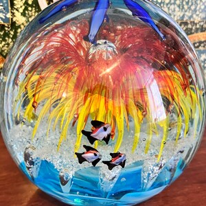 May include: A glass paperweight with a blue, yellow, and red flower inside. The flower is surrounded by small white bubbles and four small black and white fish.