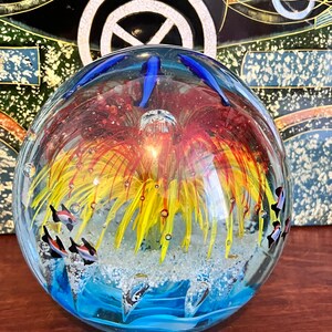 May include: A glass paperweight with a blue, white, and yellow seascape design. The paperweight features a yellow and red flower with white and black fish swimming around it.