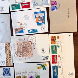 以下が含まれることがあります： 様々なデザインと色を持つ、ヴィンテージの封筒と切手のコレクション。封筒には、建物、肖像画、シンボルの画像が描かれた切手が貼られています。一部の封筒にはヘブライ語と英語のテキストがあり、郵便マークと日付が記載されています。