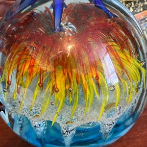May include: A glass paperweight with a blue, yellow, and red flower inside. The flower is surrounded by small bubbles. The paperweight is on a wooden surface.