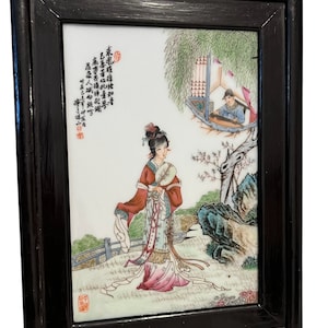 Puede incluir: Un azulejo de porcelana enmarcado con una escena pintada a mano de una mujer en un jard&iacute;n. La mujer lleva una t&uacute;nica colorida y sostiene un rollo. El azulejo est&aacute; rodeado por un marco de madera negro.