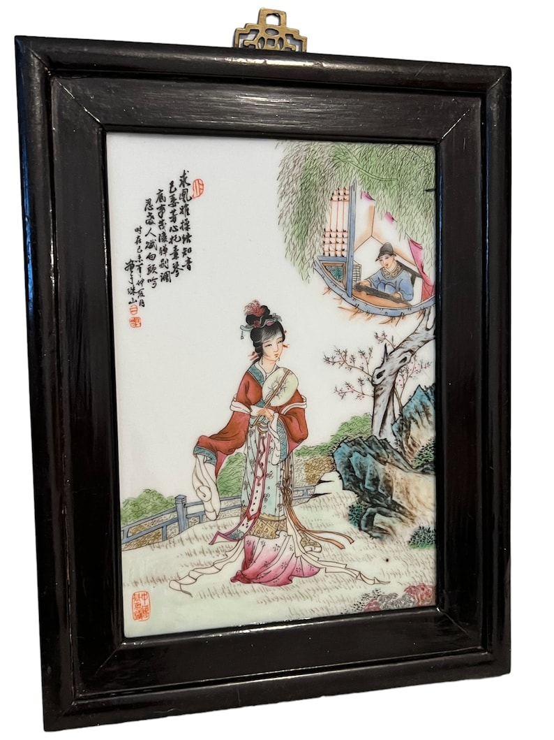 Puede incluir: Un azulejo de porcelana enmarcado con una escena pintada a mano de una mujer en un jard&iacute;n. La mujer lleva una t&uacute;nica colorida y sostiene un rollo. El azulejo est&aacute; rodeado por un marco de madera negro.