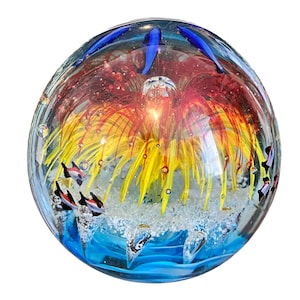 May include: A glass paperweight with a colorful, abstract design. The paperweight features a red, yellow, and orange flower-like design with blue accents. There are small, white bubbles throughout the paperweight.