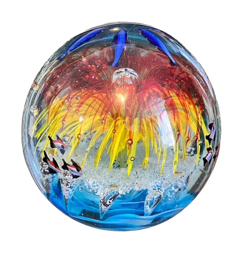 May include: A glass paperweight with a colorful, abstract design. The paperweight features a red, yellow, and orange flower-like design with blue accents. There are small, white bubbles throughout the paperweight.