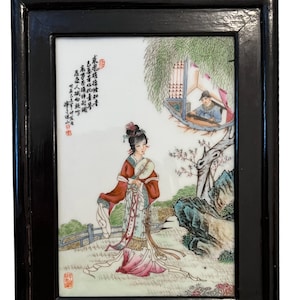 Puede incluir: Un azulejo de porcelana enmarcado con una escena pintada de una mujer en un entorno de jard&iacute;n. La mujer lleva una t&uacute;nica colorida y sostiene un abanico. La escena tambi&eacute;n incluye a un hombre tocando un instrumento musical en un edificio en el fondo. El azulejo est&aacute; enmarcado en madera negra con un gancho de suspensi&oacute;n de lat&oacute;n.