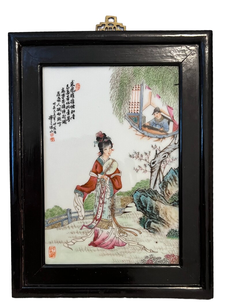 Puede incluir: Un azulejo de porcelana enmarcado con una escena pintada de una mujer en un entorno de jard&iacute;n. La mujer lleva una t&uacute;nica colorida y sostiene un abanico. La escena tambi&eacute;n incluye a un hombre tocando un instrumento musical en un edificio en el fondo. El azulejo est&aacute; enmarcado en madera negra con un gancho de suspensi&oacute;n de lat&oacute;n.