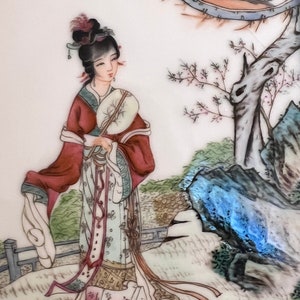 Puede incluir: Un plato de porcelana pintado a mano que representa a una mujer con una t&uacute;nica roja y blanca sosteniendo un abanico. La mujer est&aacute; de pie en un entorno de jard&iacute;n con un &aacute;rbol y rocas en el fondo. El plato est&aacute; decorado con delicados detalles florales.