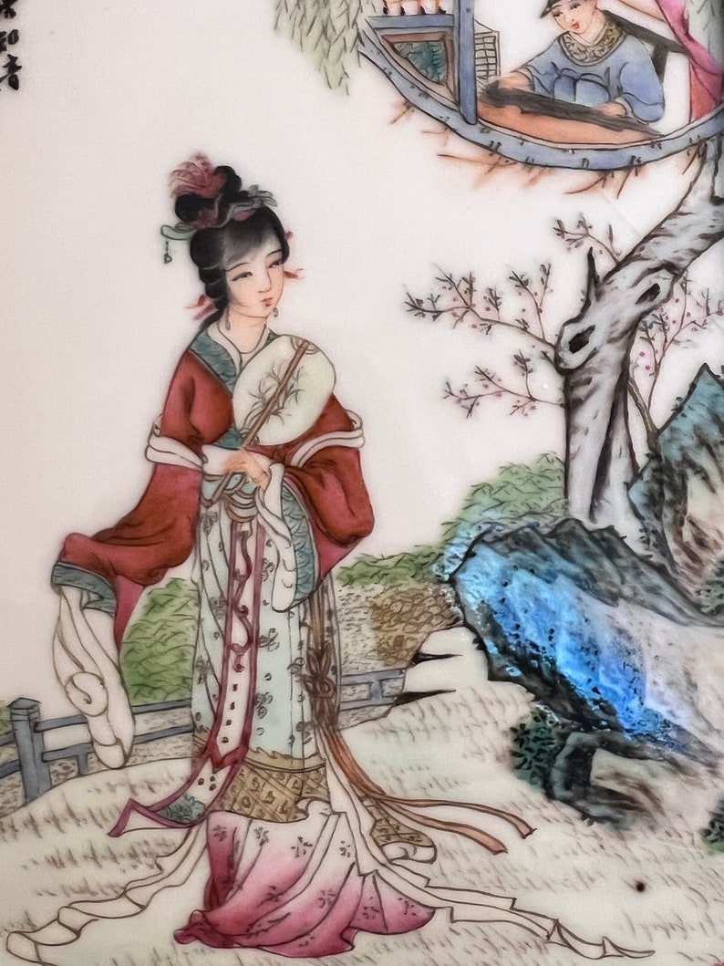 Puede incluir: Un plato de porcelana pintado a mano que representa a una mujer con una t&uacute;nica roja y blanca sosteniendo un abanico. La mujer est&aacute; de pie en un entorno de jard&iacute;n con un &aacute;rbol y rocas en el fondo. El plato est&aacute; decorado con delicados detalles florales.