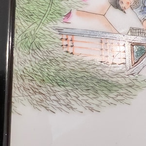 Puede incluir: Una pintura enmarcada vintage que representa una escena de una persona con una t&uacute;nica azul y blanca de pie frente a un edificio con un techo verde. La pintura est&aacute; sobre un fondo de follaje verde.