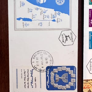 以下が含まれることがあります： 青と黒の切手が貼られた白い封筒。切手には地図、羅針盤、メノラーが描かれています。ヘブライ語のテキストが切手と封筒に表示されています。封筒には27.2.52の日付が刻印されています。