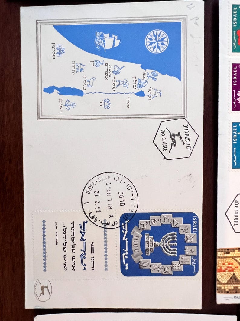 以下が含まれることがあります： 青と黒の切手が貼られた白い封筒。切手には地図、羅針盤、メノラーが描かれています。ヘブライ語のテキストが切手と封筒に表示されています。封筒には27.2.52の日付が刻印されています。