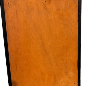 Puede incluir: La parte posterior de un cuadro enmarcado en negro con un respaldo de madera. El marco tiene dos cierres de lat&oacute;n en la parte superior.