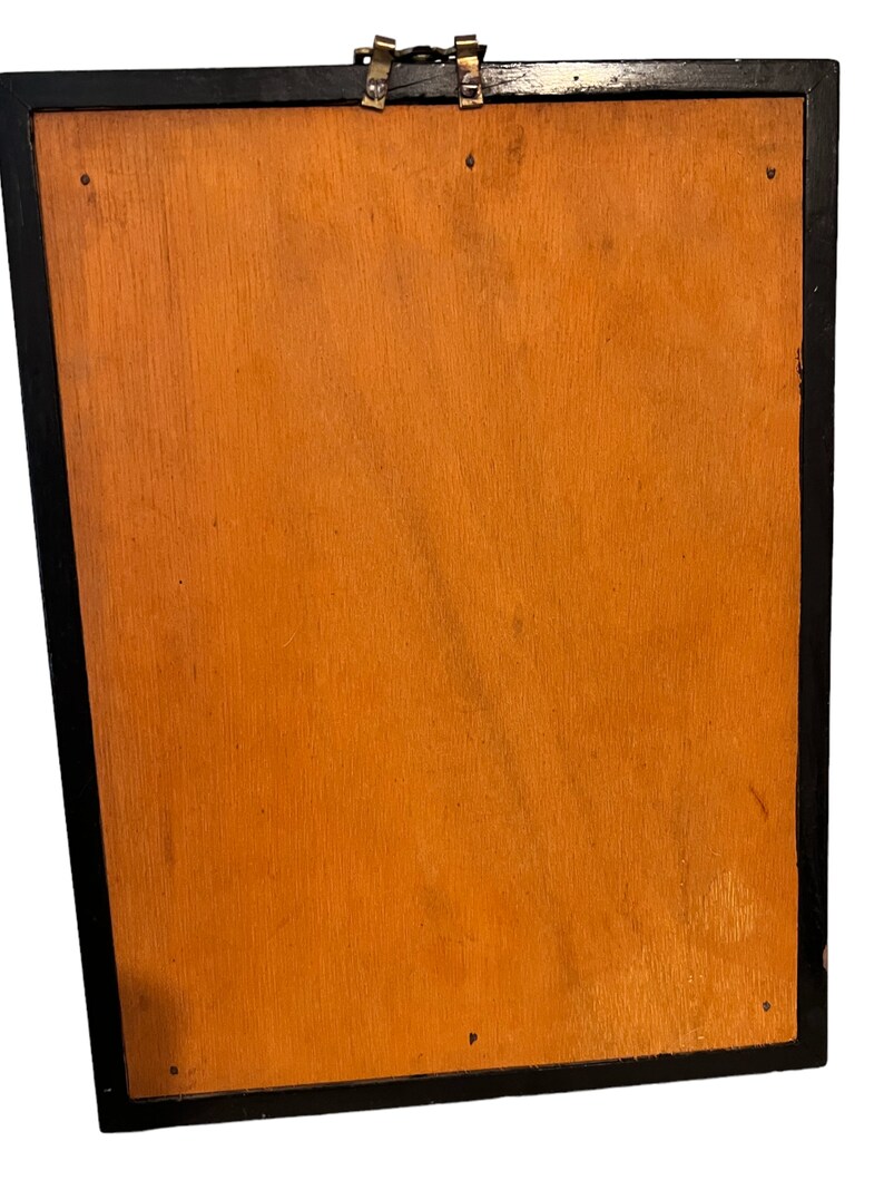 Puede incluir: La parte posterior de un cuadro enmarcado en negro con un respaldo de madera. El marco tiene dos cierres de lat&oacute;n en la parte superior.