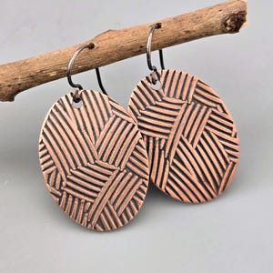 May include: A pair of copper-colored oval earrings with a geometric, woven-like pattern. The earrings are suspended from silver-colored ear wires and are displayed on a small, brown twig against a gray background. The earrings have a textured surface.