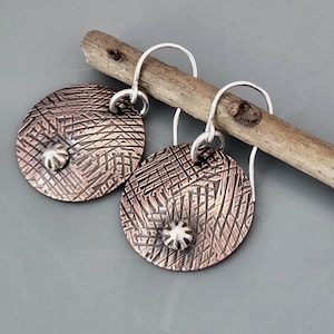 May include: A pair of handcrafted copper-colored earrings with a textured, cross-hatched design. Each earring features a small, silver-colored accent and a silver hook. The earrings are displayed on a wooden stick against a gray background.