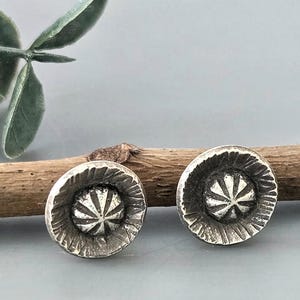 May include: A pair of silver-toned stud earrings. Each earring has a circular design with a textured outer edge and a starburst pattern in the center. The earrings are displayed on a neutral background with a wooden branch and greenery.