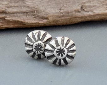 May include: A pair of round silver stud earrings featuring a sunburst pattern with radiating lines and a small star-shaped design at the center. The metal has a darkened, oxidized finish in the recessed areas to highlight the engraved details.
