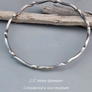 May include: A silver-colored bangle bracelet with a textured, organic design. The bracelet has a 2.5-inch inner diameter and is considered a size medium. The bracelet is displayed on a gray background with a piece of driftwood.