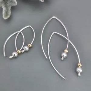 May include: Silver-toned drop earrings with a curved design. Each earring features a silver bead, a gold bead, and a smaller silver bead. The earrings are displayed on a gray background.
