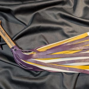 Peut inclure: Flogger artisanal avec un manche en bois et une série de lanières en cuir violet, jaune et blanc. Le manche est de couleur marron clair et possède une boucle en cuir. Les lanières sont fixées avec des rivets.