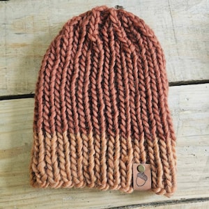 May include: A knitted beanie hat with a brown and orange ombre color scheme. The hat has a small leather patch with an acorn design on the side.